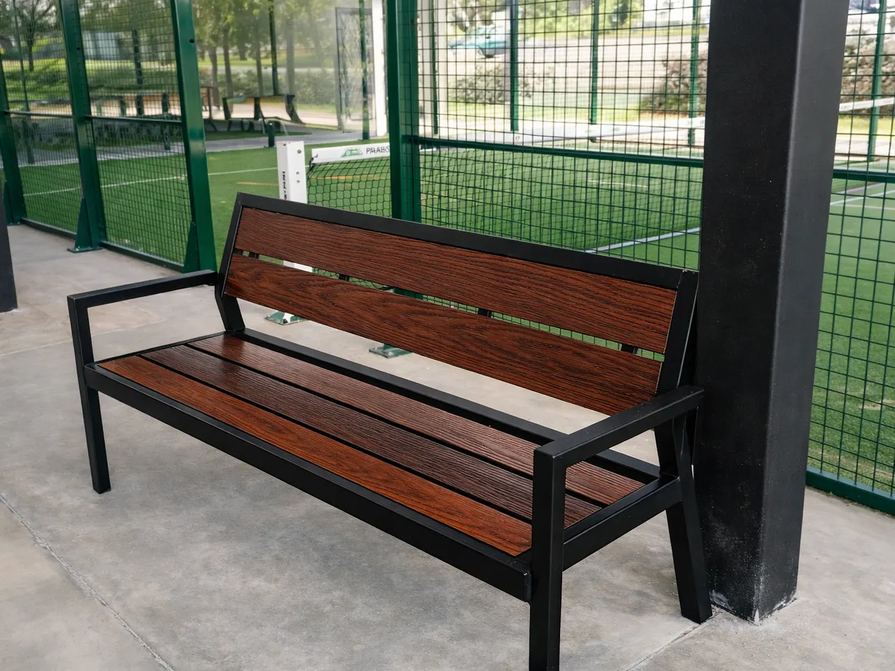 Banca con respaldo de tablillas color café frente a una cancha con malla verde