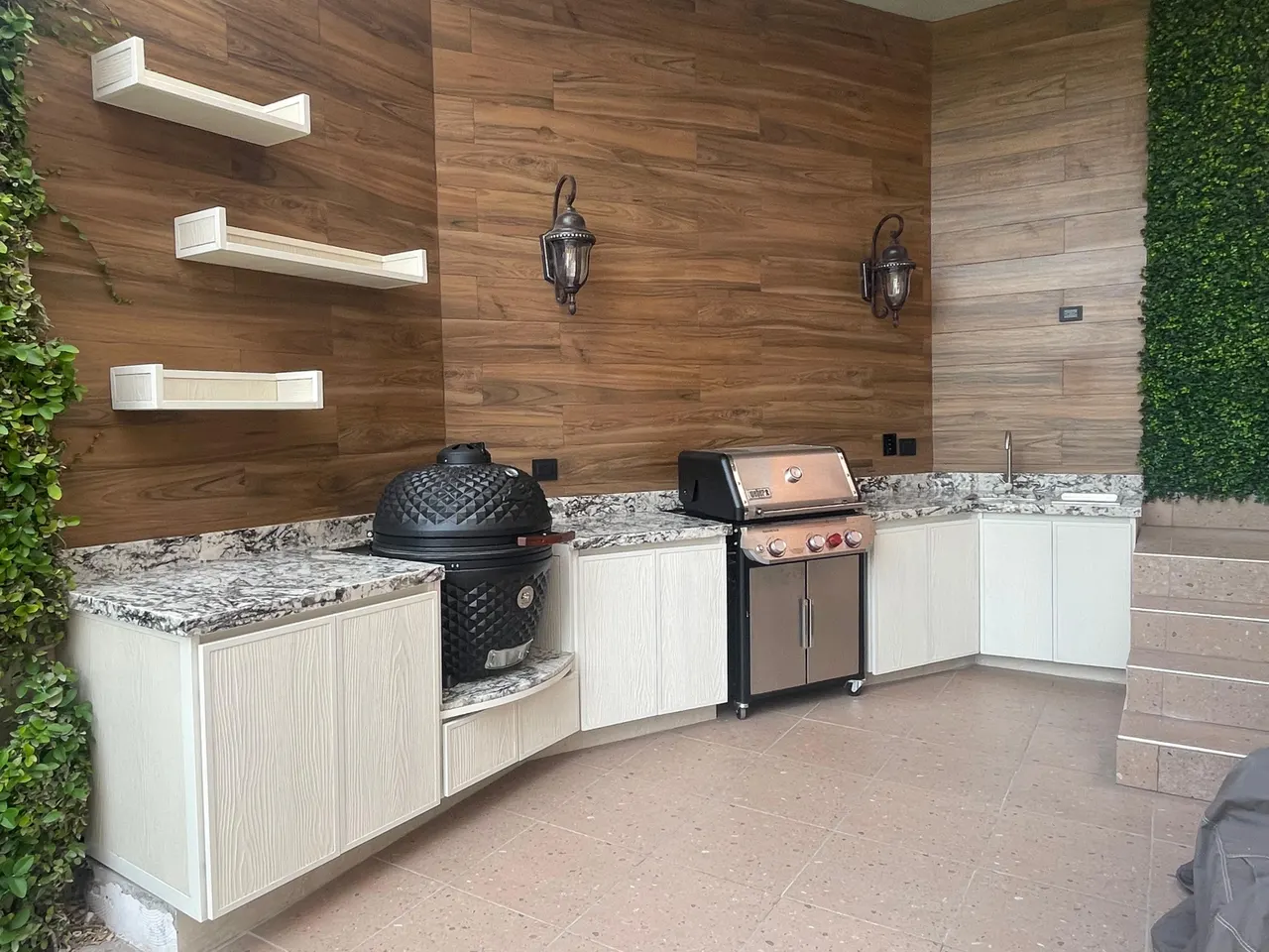 Terraza techada con una cocina exterior en escuadra, cubierta de granito, gabinetes claros, un asador tipo kamado y un asador de gas