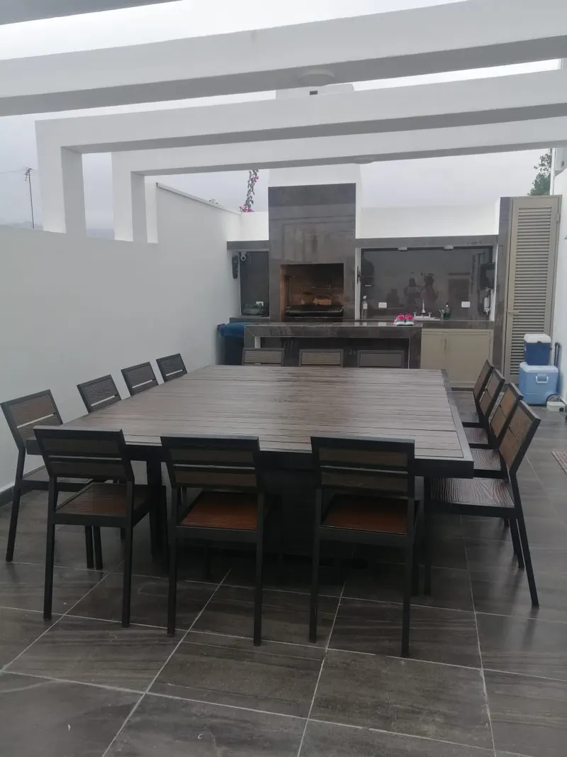 Comedor cuadrado grande con sus sillas en una terraza techada, con un asador al fondo