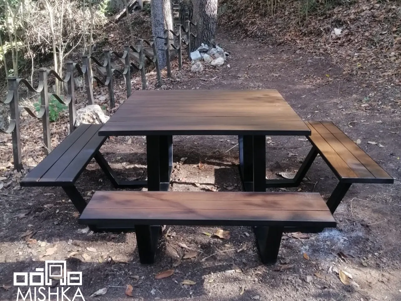 Mesa tipo picnic cuadrada con cuatro bancas, en un terreno con árboles