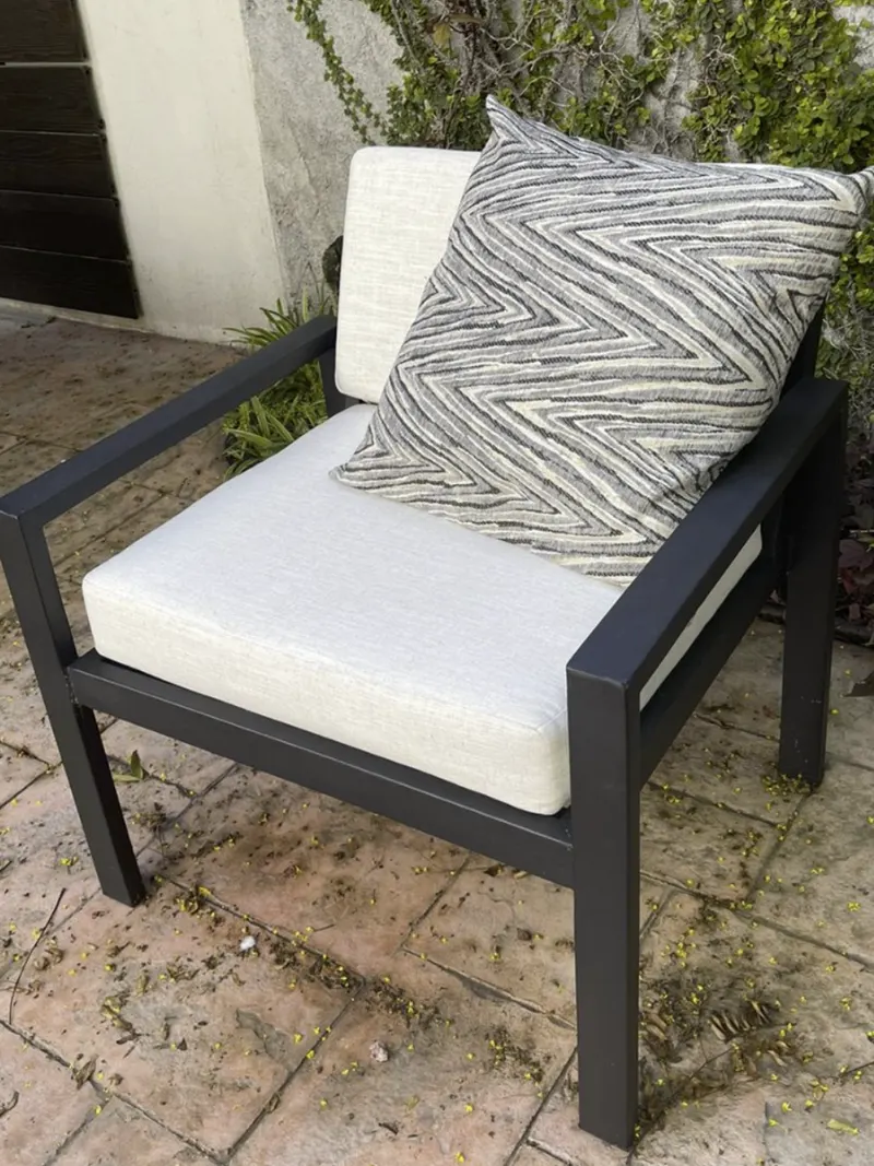 Sillón individual de estructura oscura con cojines blancos y un cojín con dibujo en zigzag, en un patio