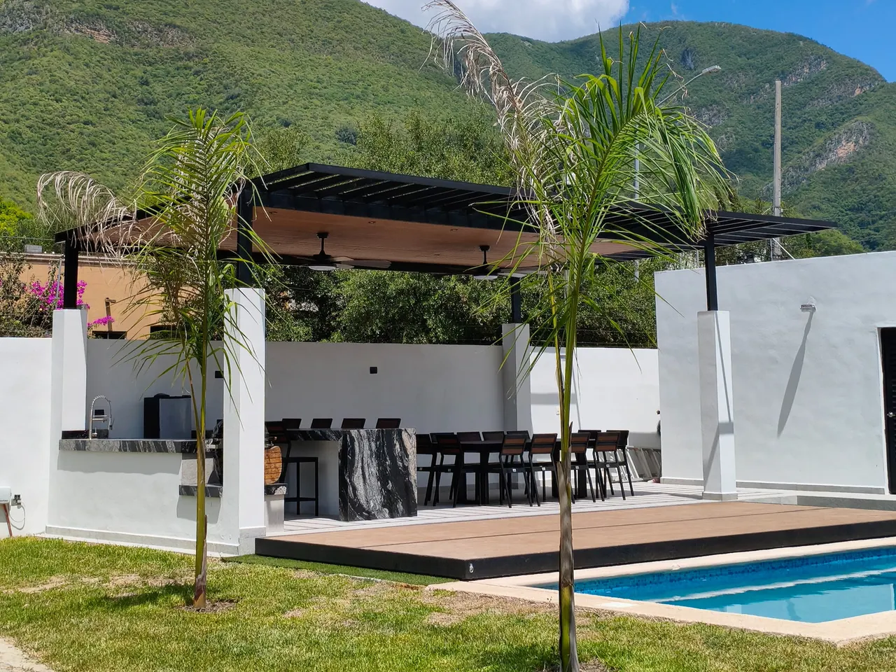 Terraza con pérgola, cocina exterior y un comedor largo, junto a una alberca y con cerros al fondo